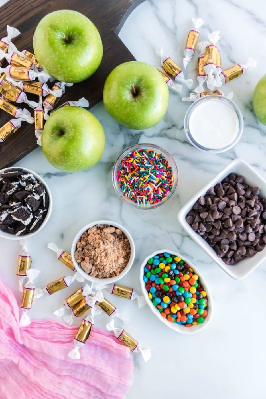 Caramel Apple ingredients on table in bowls: m&ms, chocoalte chips, sprinkles, crushed cookies, and more
