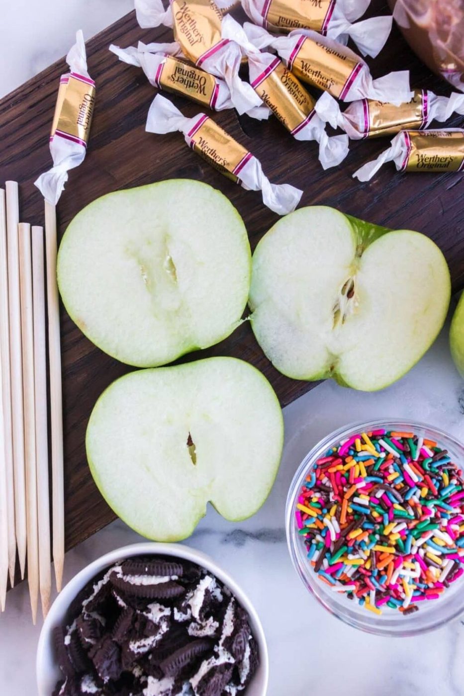 sliced apples on counter with wrapped caramels, and bowls of crushed cookies and sprinkles beside the apples