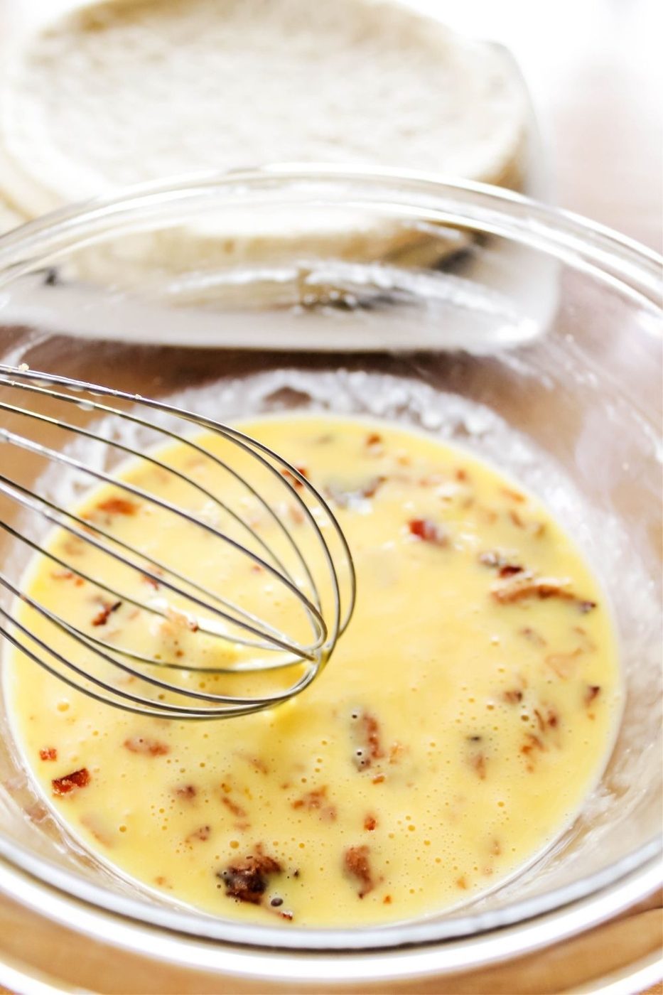 eggs whisked in a bowl with bacon and pizza crust on counter behind it