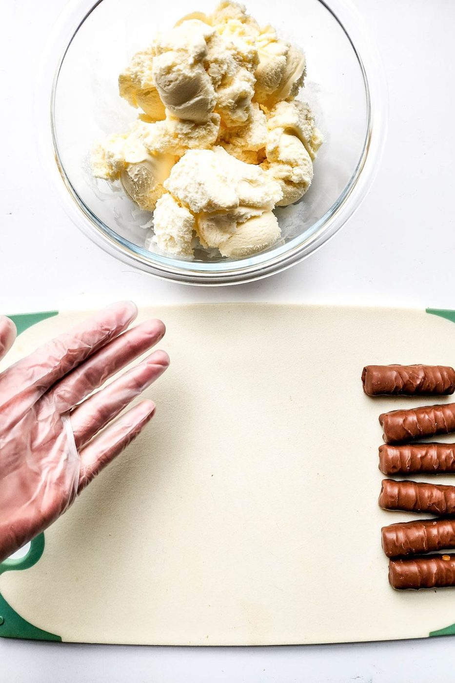ice cream on cutting board with Twix bars by it and scoops of ice cream in a bowl above it on counter 