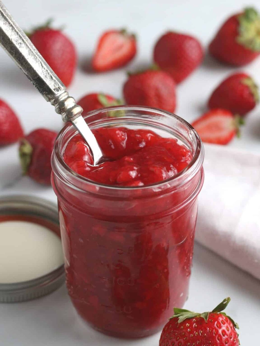 Strawberry Sauce in a glass jar with strawberries behind it on the table