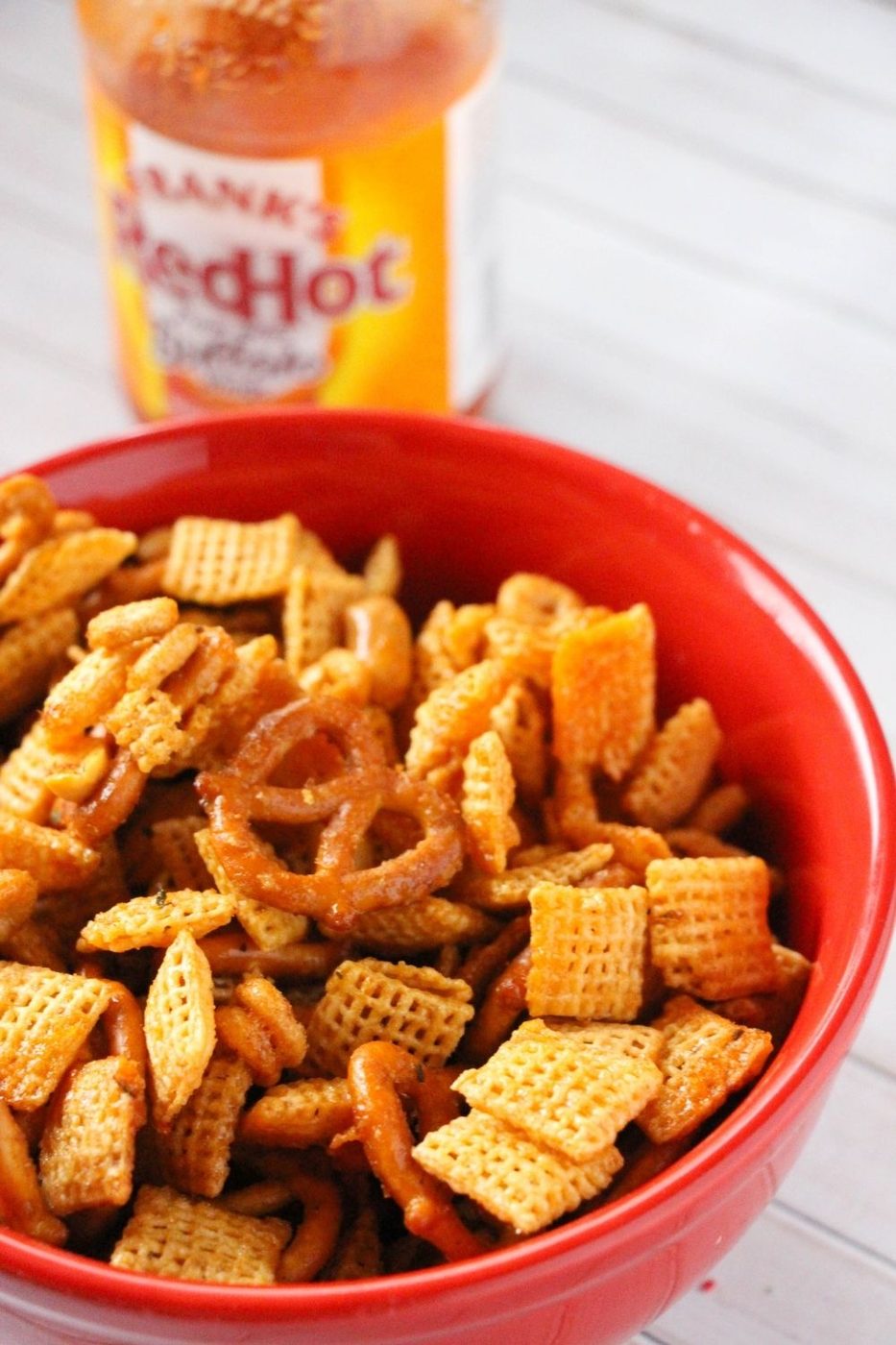 party mix in a red bowl with hot sauce on table behind it