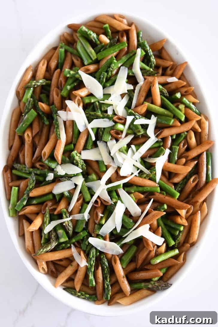 Top-down view of penne pasta in a white serving dish, generously adorned with roasted asparagus, balsamic butter, and Parmesan cheese.