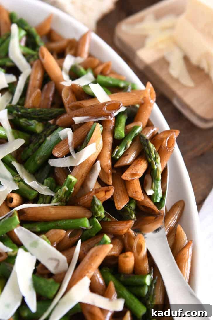 A serving spoon in a white dish filled with balsamic penne pasta, roasted asparagus, and a generous sprinkle of Parmesan cheese.