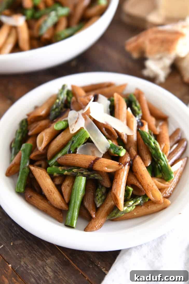 A white plate showcasing balsamic-drenched penne pasta, roasted asparagus, and a topping of Parmesan cheese, ready to be enjoyed.