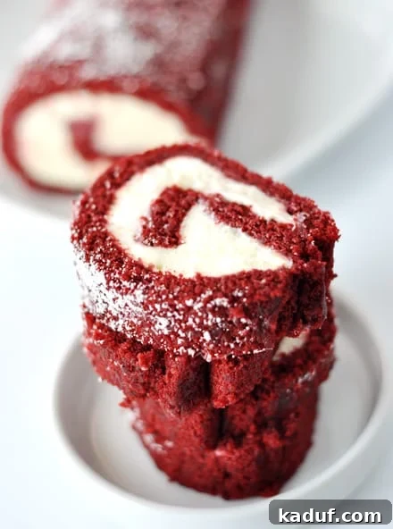 Crimson Velvet Swirl Cake 3 Slices of a cream cheese filled red velvet spiraled cake roll stacked on a white plate, with the remaining roll behind it, showing its elegant spiral design.