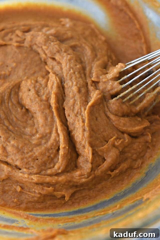 Pumpkin Donut Muffin Bites 5 A hand gently whisking pumpkin muffin batter in a clear glass bowl, ensuring no dry streaks remain.