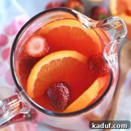 Overhead view of a glass pitcher filled with vibrant red party punch, elegantly garnished with fresh orange slices and whole strawberries, ready for serving.