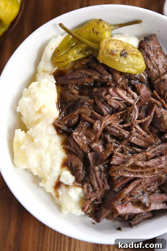 Homemade Mississippi Pot Roast 6 Top-down view of a white shallow bowl featuring mashed potatoes, generously shredded Mississippi pot roast with gravy, and two whole pepperoncini for garnish.