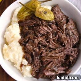 White shallow bowl with mashed potatoes, two pepperoncini, and shredded beef.