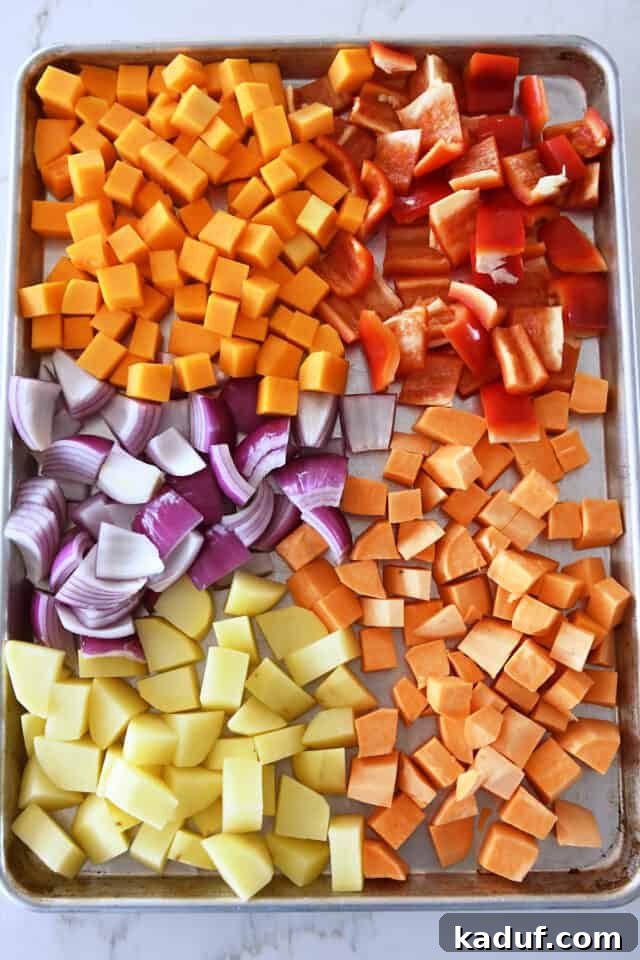 Hearty Balsamic Glazed Root Vegetables 3 An array of uncooked butternut squash, sweet potatoes, red bell peppers, red onion, and Yukon Gold potatoes, chopped and ready on a sheet pan.