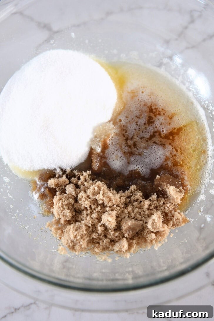 Granulated sugar, brown sugar, and warm brown butter are mixed in a clear glass bowl, forming the flavorful base.