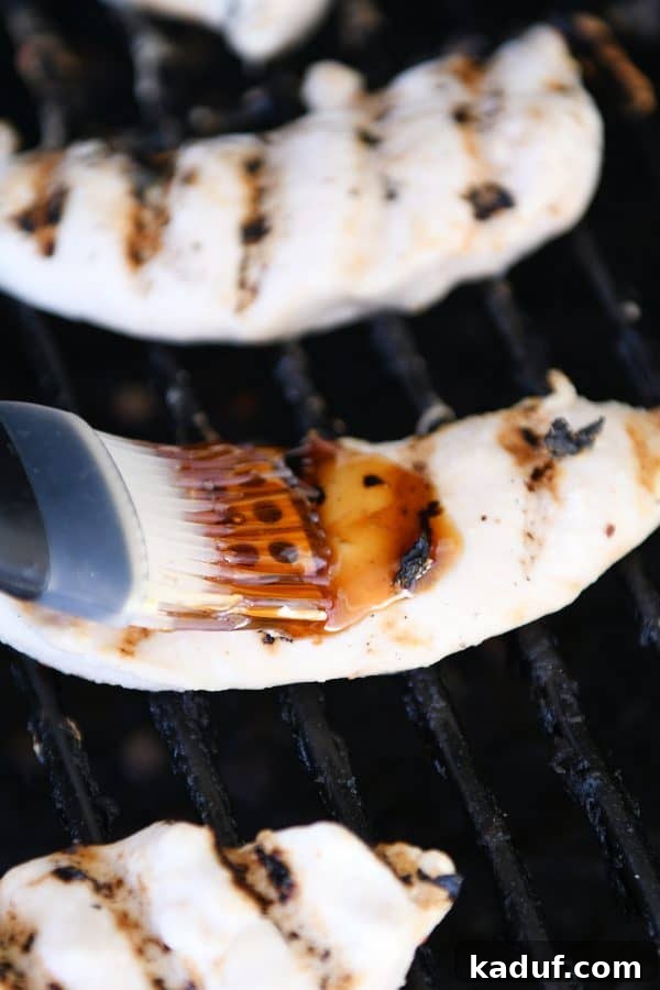 Caramel Coconut Chicken 9 A chef's hand using a silicone basting brush to generously coat pieces of grilled coconut chicken with a sticky glaze directly on the grill grates. The chicken has visible char marks and a tempting sheen.