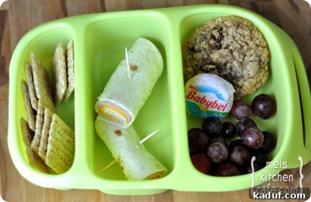Bento Box Lunch Idea 1 Top view of a green divided lunch tray featuring reduced-fat Triscuit crackers, a ham and cheese roll-up, fresh grapes, a Babybel cheese, and a homemade chocolate chip cookie, demonstrating a healthy kid's lunch.
