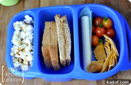 Bento Box Lunch Idea 2 Top view of a blue divided lunch box with kettle corn, a PB&H sandwich on homemade bread, string cheese, fresh cherry tomatoes, and dried mangos, showcasing a balanced and appealing lunch for kids.