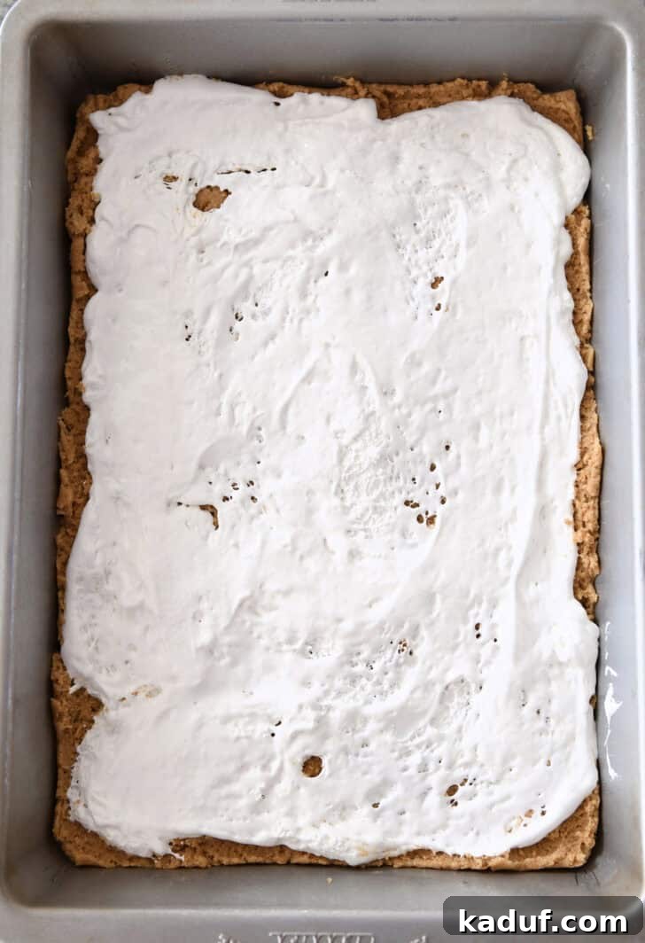 Irresistible S'mores Bars 5 Top-down view of a metal baking pan lined with a graham cracker cookie dough base, topped with an evenly spread layer of gooey marshmallow creme. This shows the second layer of the s'mores bars.