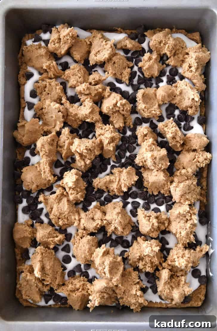 Irresistible S'mores Bars 7 Top-down view of a metal baking pan with dollops of the remaining graham cracker cookie dough scattered over the chocolate chips and marshmallow layer. This demonstrates the final top layer of the s'mores bars before baking.