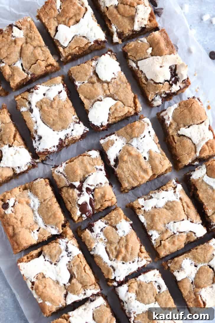 Irresistible S'mores Bars 9 Top-down view of golden-brown s'mores cookie bars, freshly cut into squares, with prominent fissures of gooey marshmallow and melted chocolate visible throughout each piece, indicating their rich and soft texture.