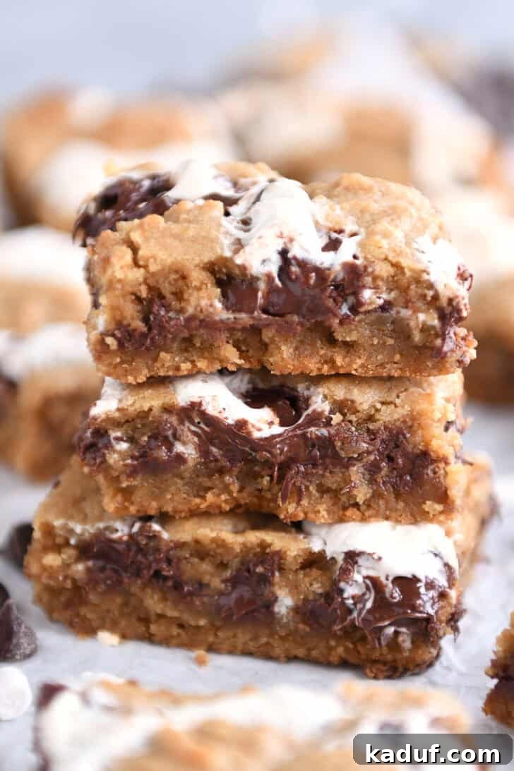 Irresistible S'mores Bars 10 Three s'mores cookie bars are neatly stacked on top of each other on a piece of parchment paper, highlighting their golden-brown edges, gooey interiors, and rich layers.