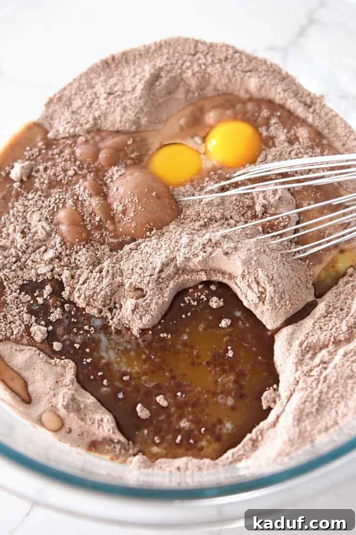 Eggs and melted butter being added to the chocolate cake mix in a clear glass bowl, preparing for the wet ingredient incorporation.