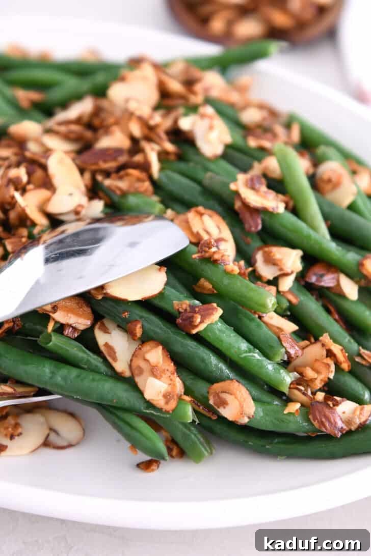 Simple Green Beans Almondine 8 Tongs grabbing a serving of perfectly steamed green beans with toasted almonds and garlic from a white serving dish.
