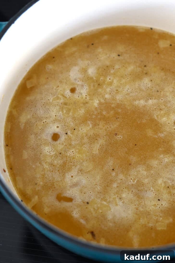 Ultimate Rice Pilaf 5 Rich chicken broth being poured into a cast iron pot with rice and orzo pilaf ingredients.