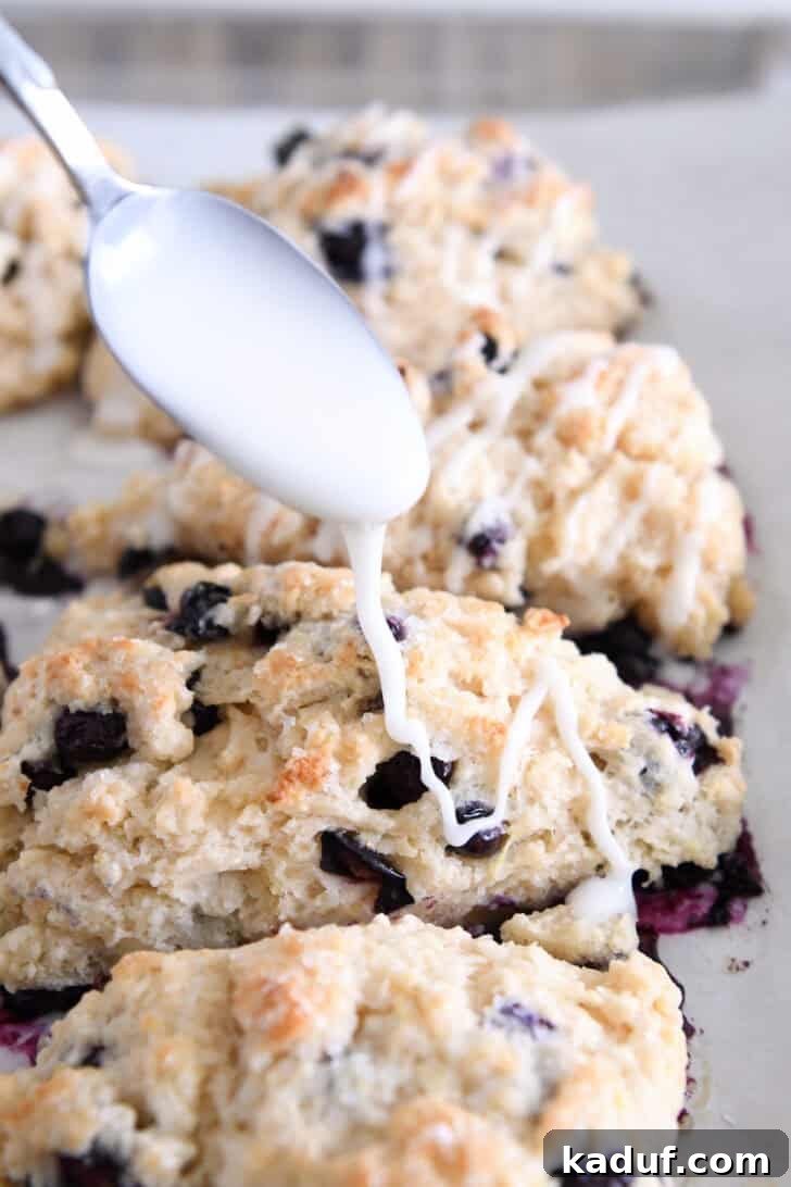 Irresistible Lemon Blueberry Scones 9 Fresh lemon glaze being drizzled over warm, baked lemon blueberry scones on a baking sheet.