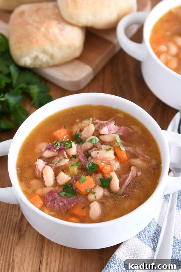 Hearty Slow Cooked Ham and Bean Soup 2 A white bowl with handles filled to the brim with slow cooker ham and bean soup, garnished with fresh parsley.