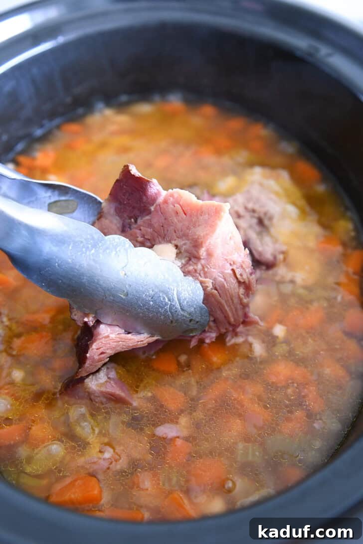 Hearty Slow Cooked Ham and Bean Soup 5 Tongs lifting a meaty ham bone out of a slow cooker, revealing the cooked white beans, broth, and vegetables.