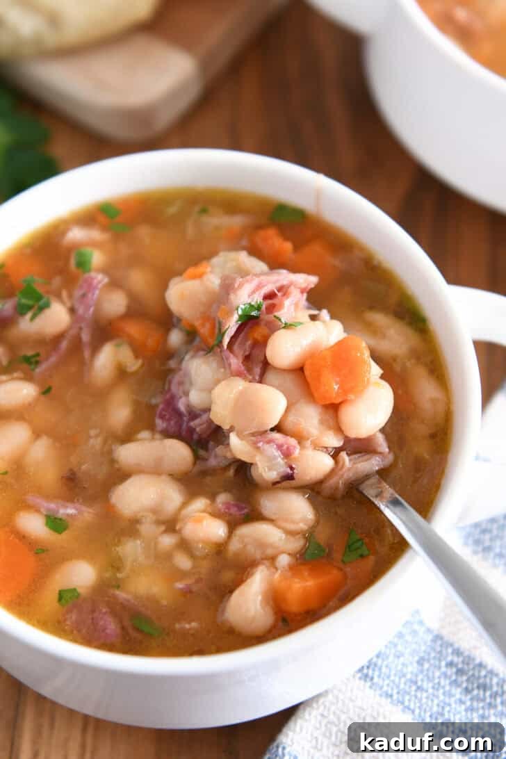 Hearty Slow Cooked Ham and Bean Soup 8 Top-down view of a white bowl with handles, overflowing with rich ham and bean soup. A spoon is lifting a perfect bite of creamy white beans, tender carrots, and succulent ham pieces.