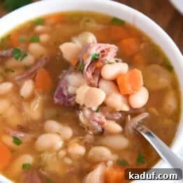 White bowl with handles with spoon lifting bite of white beans, carrots and ham.