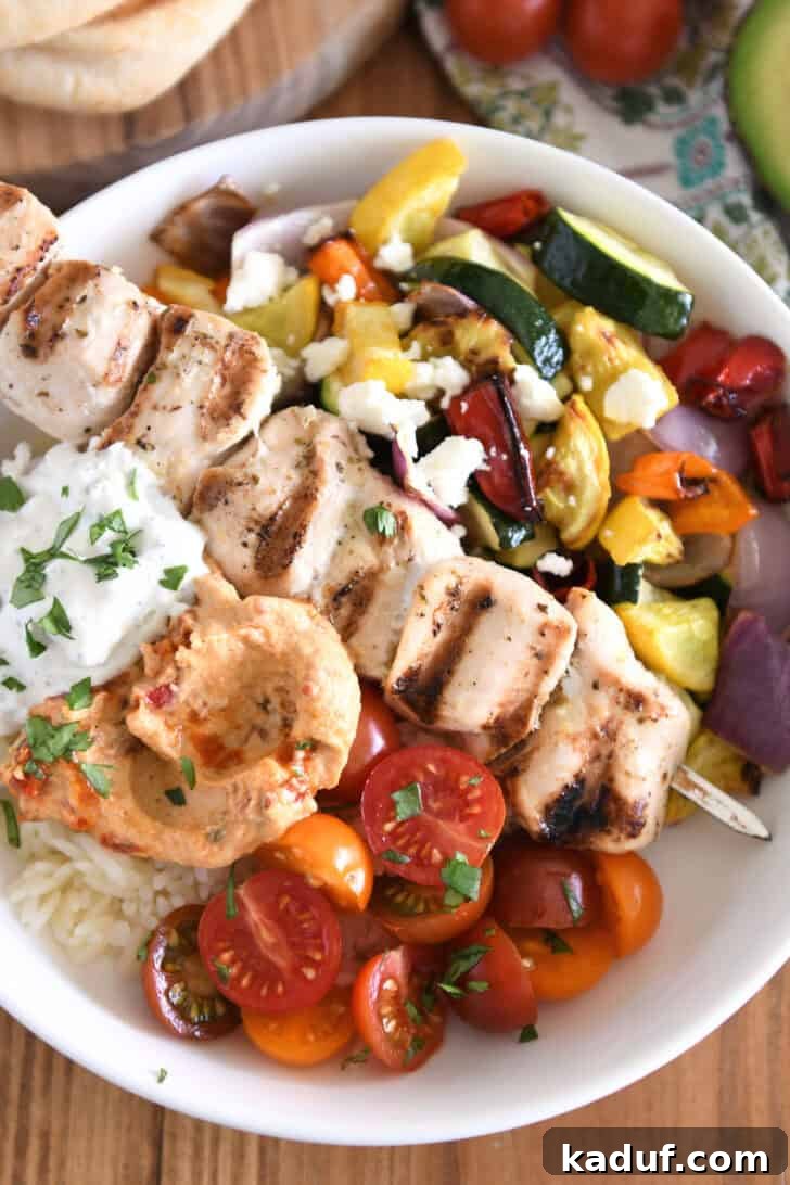 A vibrant overhead shot of a white bowl filled with tantalizing Chicken Souvlaki. It features perfectly grilled lemon-oregano chicken skewers, tender roasted zucchini, bell peppers, and red onions, alongside juicy cherry tomatoes. The bowl is beautifully rounded out with fluffy garlic rice, a generous dollop of creamy hummus, and a swirl of refreshing tzatziki sauce, creating a harmonious blend of Mediterranean flavors.