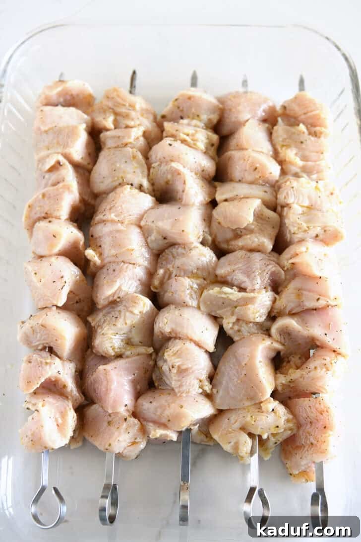 Raw chicken pieces, perfectly marinated in lemon and herb mixture, expertly threaded onto metal skewers and arranged in a glass 9x13-inch baking dish, ready for grilling.