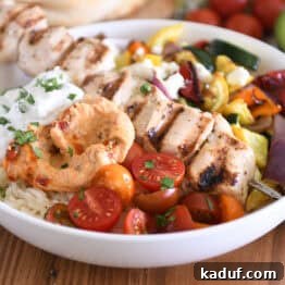 White bowl with metal skewer with chicken pieces, roasted squash, zucchini, red onions and bell peppers, cherry tomatoes, rice, hummus and tzatziki sauce.