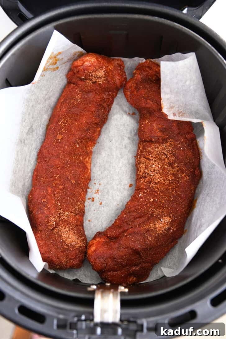 Quick and Tender Air Fryer Pork Tenderloin 5 Two spice-crusted uncooked pork tenderloins arranged on parchment paper inside an air fryer basket, ready for cooking.