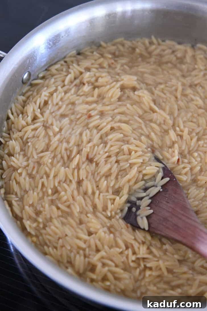 Effortless Tuna Zucchini Orzo Skillet 4 Orzo pasta gently simmering in chicken broth within a stainless steel skillet, indicating the liquid absorption phase of the one-pan cooking.