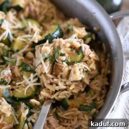 Serving spoon scooping serving of cooked orzo, tuna, zucchini and Parmesan in stainless skillet.