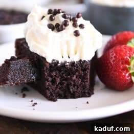 Piece of chocolate olive oil cake topped with whipped cream and crispy chocolate pearls with bite of cake on fork on white plate.