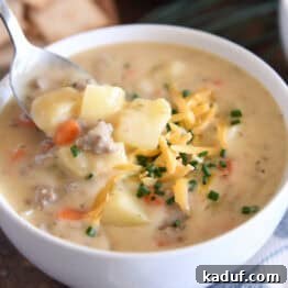White bowl with spoon scooping ingredients in creamy cheesy soup with cooked potatoes, ground beef, and carrots and topped with shredded cheddar cheese and chopped chives.