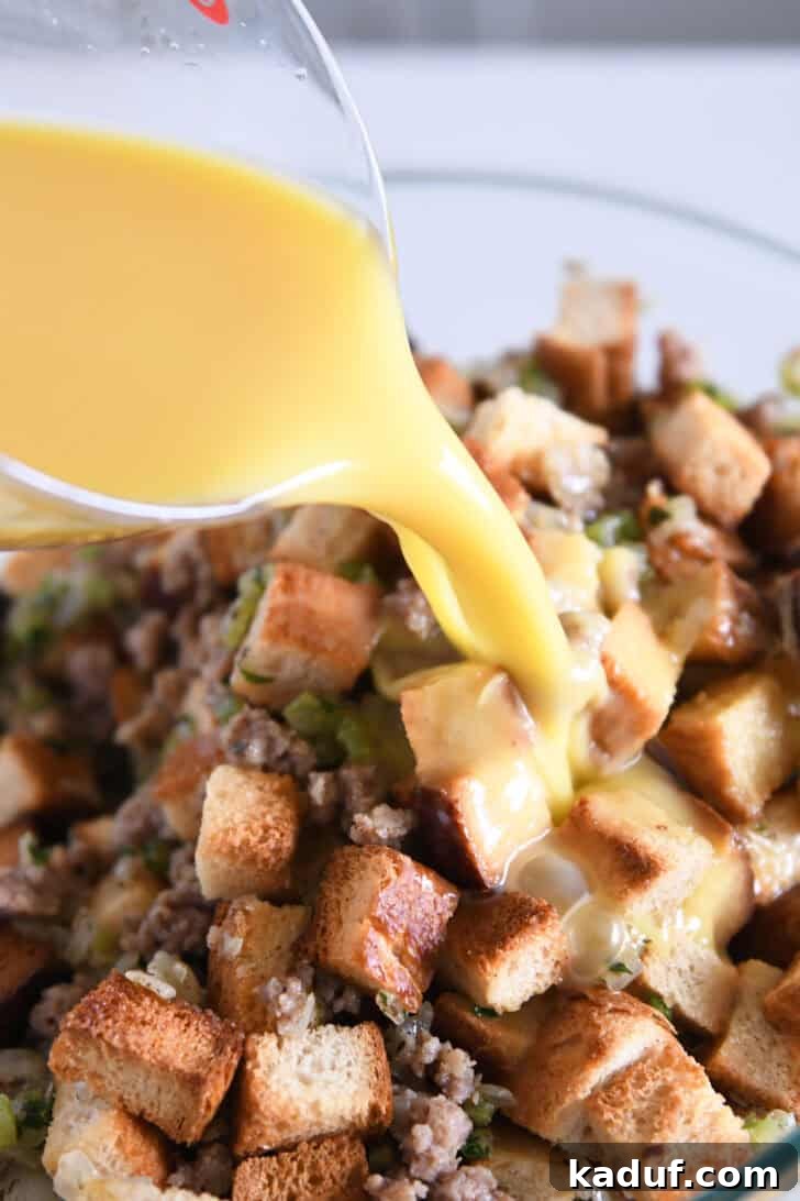 Hearty Sausage and Herb Stuffing 5 Hands pouring whisked eggs and chicken broth over a mixture of toasted bread, cooked sausage, and vegetables in a glass bowl.
