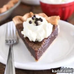 Triangle slice of chocolate chess pie on white plate with fork to the side and a dollop of whipped cream on top of pie topped with chocolate crisp pearls.