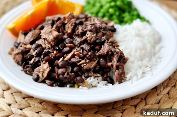 Shredded pork and black beans next to cooked white rice, peas, and orange slices on a white plate, showcasing a complete and inviting meal.