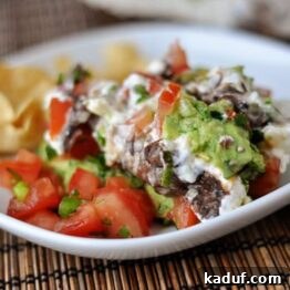 White plate with a big serving of 7-layer dip and tortilla chips on the side.