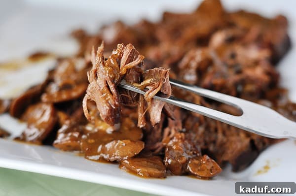 Fork Tender Brisket Braised with Forest Mushrooms 3 Close-up of shredded braised brisket on a white platter with a fork lifting a bite, showcasing its tenderness.