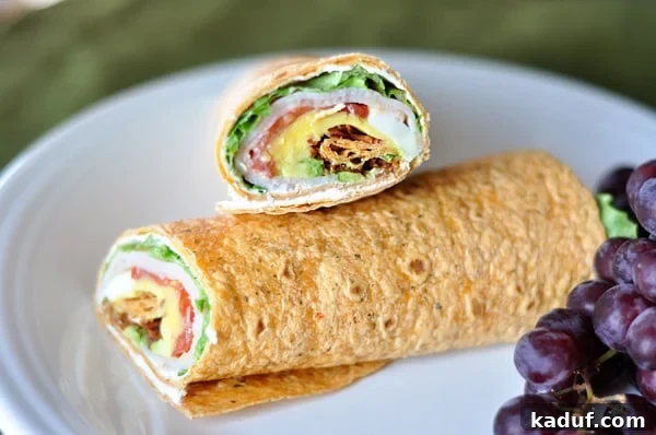 Smoky Cobb Turkey Handhelds 2 A vibrant smoked turkey cobb wrap, sliced in half, revealing layers of fresh ingredients like lettuce, tomato, smoked turkey, hard-boiled egg, avocado, and bacon, served on a white plate alongside purple grapes.