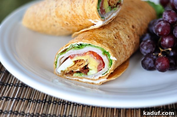 Smoky Cobb Turkey Handhelds 3 A close-up view of the interior of a smoked turkey cobb wrap, cut diagonally, showcasing the tightly packed layers of turkey, bacon, egg, avocado, tomato, and lettuce, on a white plate.