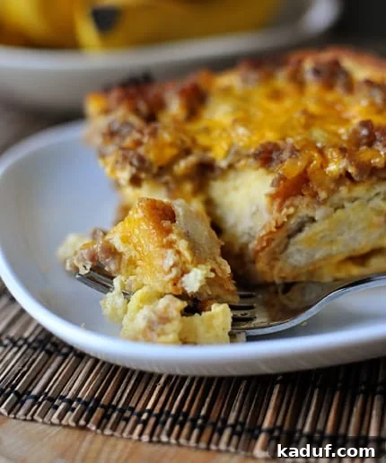 Overnight Sausage Egg Casserole 2 A delectable piece of Make-Ahead Sausage and Egg Breakfast Bake on a pristine white plate, with a fork poised to take a bite, showcasing its golden-brown top and fluffy interior.
