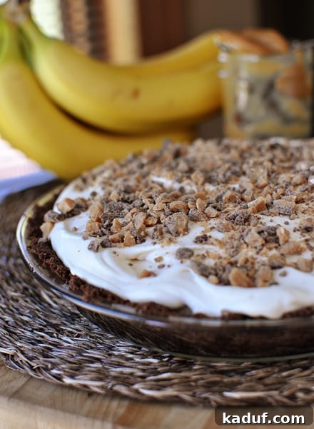 Caramelized Banana Dream Pie 2 A chocolate crust banana pie with whipped cream and chopped toffee bits in a glass pie dish and bananas in the background.