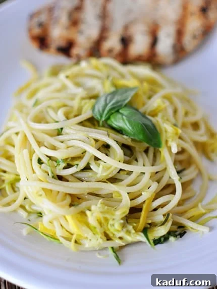 Garden Fresh Zucchini and Yellow Squash Noodles 3 Close-up of cooked pasta with julienned zucchini and yellow squash, garnished with fresh basil leaves and a grilled chicken breast in the background.