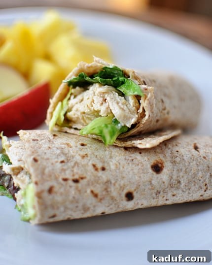 Ultimate Caesar Chicken Wraps 2 A chicken caesar salad wrap cut in half on a white plate with fruit on the background.